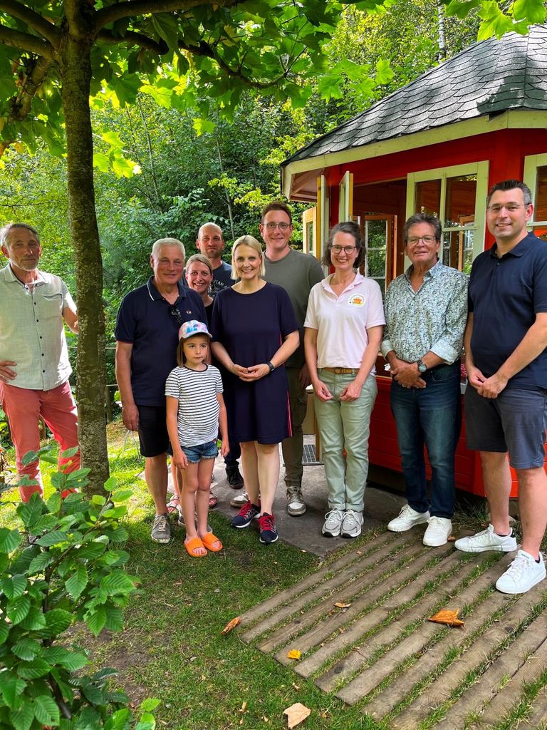 Gruppe von Menschen vor rotem Gartenhäuschen im Grünen, fröhlich posierend.